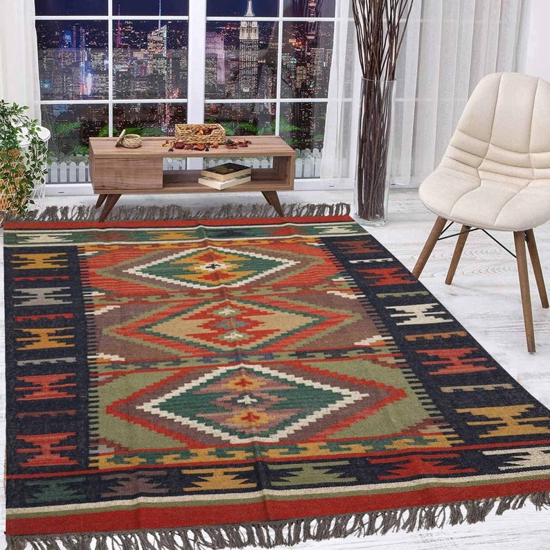 May include: A rectangular, patterned rug with a geometric design in shades of red, green, brown, and black. The rug has a fringed edge and is placed on a light-colored floor. The rug is in a room with a window and a chair.