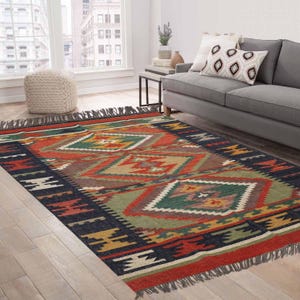 Op de afbeelding: Een rechthoekig vloerkleed met een geometrisch patroon in rood, groen, geel en bruin. Het vloerkleed heeft een donkerblauwe rand en gefranjerde randen. Het vloerkleed ligt op een houten vloer in een woonkamer.