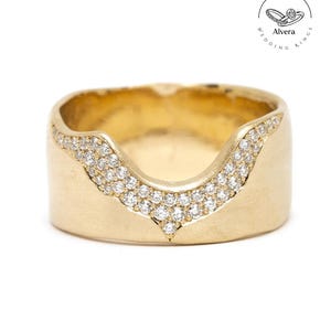 Puede incluir: Un anillo ancho de color dorado con un diseño en forma de V. El anillo está incrustado con pequeñas piedras preciosas transparentes a lo largo de la forma de V. El anillo tiene un acabado mate y una banda lisa y redondeada. Es una alianza.