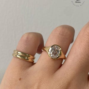 Może przedstawiać: Wyświetlane są dwa złote pierścionki. Jeden pierścionek ma duży, przezroczysty kamień o owalnym szlifie osadzony w złotej oprawie. Drugi pierścionek to prosta, polerowana złota obrączka. Pierścionki są na palcu osoby. Marka "Alvera Wedding Rings" jest w tle.