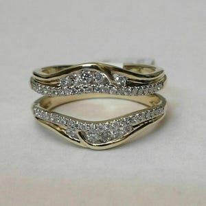 Op de afbeelding: Een gouden ringbeschermer bestaande uit twee gebogen banden die aan de zijkanten met elkaar verbonden zijn. Elke band is ingelegd met een rij kleine, heldere, rondgeslepen stenen die de golvende contouren van het metaal volgen.