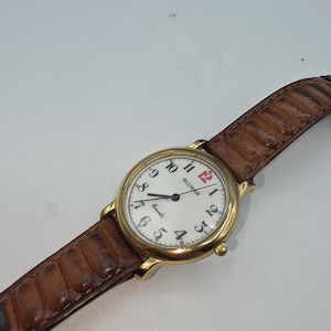 May include: A gold-toned wristwatch with a white face and black numerals. The watch has a brown leather band. The watch face reads "BLUMAR" and "Quartz". The number 12 is in red.