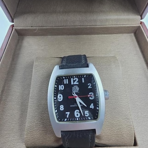 May include: A rectangular silver wristwatch with a black face and a black leather strap. The watch face has white numerals and hands, with a lion emblem. The watch is displayed in a brown presentation box.