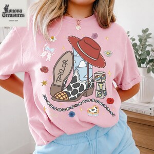 A woman wearing a pink t-shirt with a cartoon cowboy shoe and various cartoon-style elements, such as a red hat, flowers, and stars, along with the text "I know you're a girl".