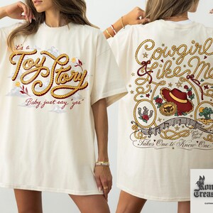 two white t-shirts with a cowboy theme. The t-shirt on the left has a red and gold design with the text "It's a Toy Story Baby just say 'yep'" and a cowboy hat. The t-shirt on the right has a red and gold design with the text "Cowsgirl Like Me Takes One to Know One" and a cowboy hat.