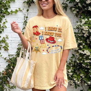A woman wearing a yellow t-shirt with various cartoon-style illustrations and text, standing in front of a white brick wall with greenery.