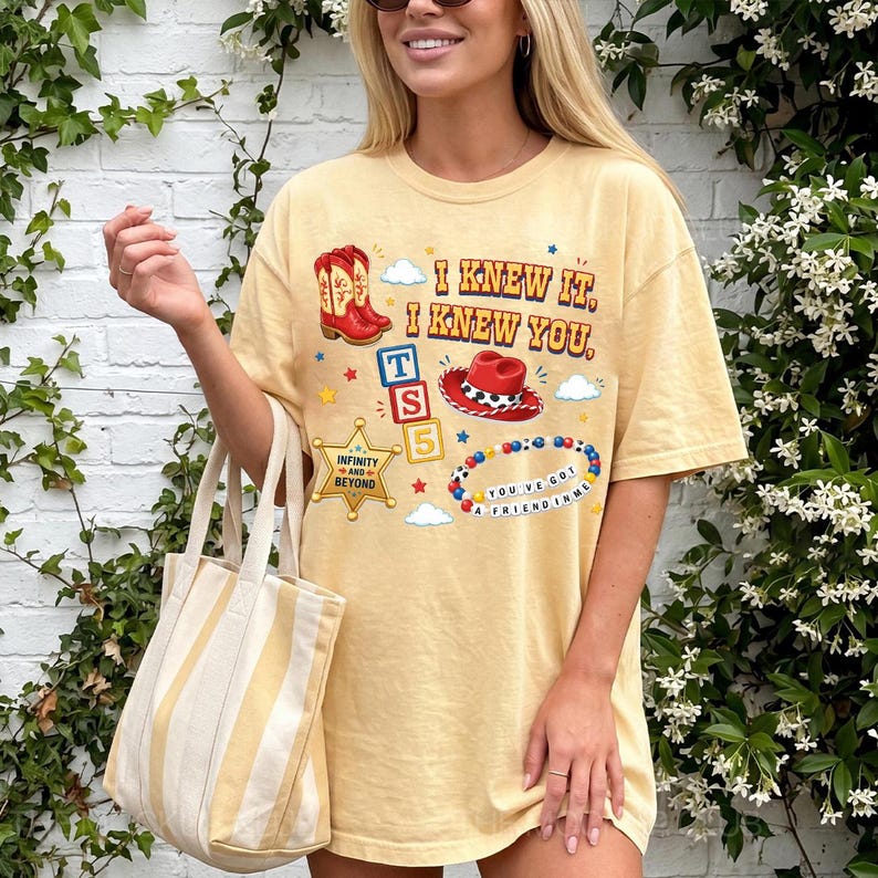 A woman wearing a yellow t-shirt with various cartoon-style illustrations and text, standing in front of a white brick wall with greenery.
