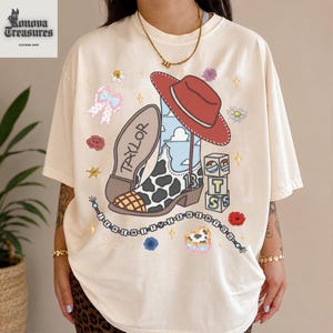A person wearing a white t-shirt with a colorful graphic design featuring a cowboy hat, boots, and various other cartoon-style elements.