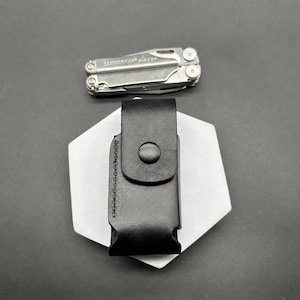 May include: A black leather sheath with a snap button closure and visible side stitching, positioned vertically. Above the sheath lies a silver-coloured Leatherman Wave multi-tool in a folded position, displaying the brand name engraved on its metal surface.