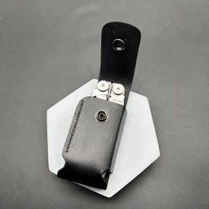 May include: A black leather sheath with a vertical flap and a metal snap closure. The sheath contains a silver multi-tool with visible circular pivot points and metallic components. The leather features visible stitching along the side edge.