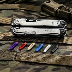 Könnte beinhalten: Ein silbernes und schwarzes Multitool mit dem Text "LEATHERMAN ARC" liegt auf einem Tarnmuster-Hintergrund. Darunter befinden sich farbige Ersatzbits in Lila, Rot, Beige, Blau, Weiß und Schwarz.