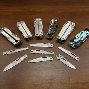 Könnte beinhalten: Eine Sammlung von Multitools und Klingen, die auf einer dunklen Holzoberfläche angeordnet sind. Die Multitools sind silber, schwarz und blau. Die Klingen haben ein gemustertes Design. Die Werkzeuge sind für verschiedene Aufgaben konzipiert.