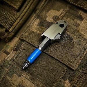 May include: A multi-functional tool featuring a metallic body with a circular hole at the top and a hexagonal shaft. The tool includes a replaceable screwdriver bit with a bright blue sleeve at the base, designed for manual fastening tasks.