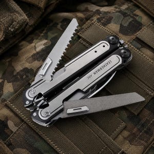 May include: A multi-tool with a silver and black finish, featuring a saw blade, knife, and other tools. The text "LEATHERMAN ARC" is visible on the handle. The tool is displayed on a camouflage background.