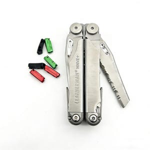 May include: A silver Leatherman Wave+ multi-tool with a partially serrated blade is displayed. The tool is open, revealing the blade. Beside it are four colored bit holders in black, green, and red.