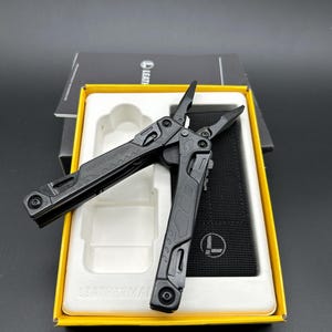 Puede incluir: Multiherramienta negra con alicates, cuchillo y otras herramientas, presentada en una caja amarilla y blanca. La herramienta está abierta, mostrando los alicates. La caja tiene el nombre de la marca "LEATHERMAN".