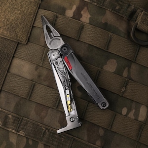 May include: A silver and black Leatherman Surge multi-tool with needle-nose pliers, a gold-colored screwdriver bit, and a red accent piece on the handle. The tool features a metallic frame with a skeletal design and is partially unfolded.