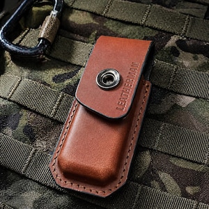 May include: A brown leather multi-tool sheath with the word "LEATHERMAN" embossed on the flap. The sheath has a snap closure and is stitched with brown thread. The background is a camouflage pattern.
