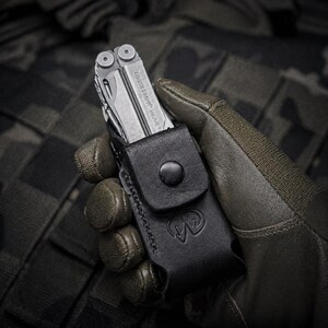 May include: A silver multi-tool with the text "LEATHERMAN WAVE+" is partially visible in a black leather sheath. The sheath has a snap closure and the Leatherman logo. The multi-tool is held in a gloved hand.