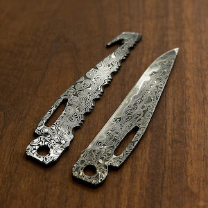 Puede incluir: Dos cuchillos de metal con un diseño de patrón único. Uno tiene un borde dentado y un gancho, el otro una hoja puntiaguda. Ambos cuchillos tienen un agujero cerca del mango y están sobre una superficie de madera.