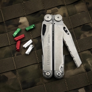 May include: A Leatherman Wave+ multi-tool with a partially serrated blade and several colored bit drivers. The multi-tool is silver and has the brand name engraved on the side. The bit drivers are red, green, and white.