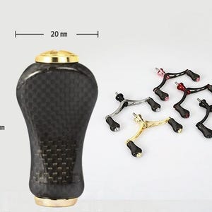 May include: A black carbon fiber fishing reel handle knob with gold-colored metal caps at the top and bottom, measuring 20 mm in width and 38 mm in height. Beside it are five additional fishing reel handles in various metallic finishes.