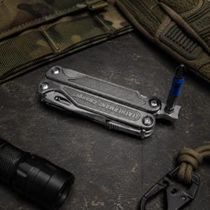 May include: A silver Leatherman Charge multi-tool in a folded position with a blue and silver screwdriver bit inserted into the tool head. The metal body features textured grip panels and visible screws, showcasing a compact and functional design.