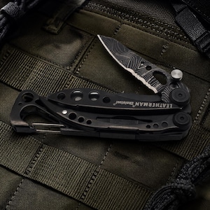 May include: Black Leatherman Skeletool multi-tool with a partially open blade. The blade has a unique pattern. The tool includes a carabiner clip and the brand name is visible. The multi-tool is on a green textured surface.