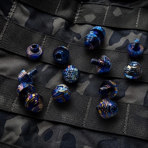 Op de afbeelding: Een assortiment blauwe en gouden metalen kralen in verschillende vormen en maten. De kralen hebben een textuur, wervelend patroon en zijn gerangschikt op een donkere camouflage achtergrond. Sommige kralen hebben schroefdraad.