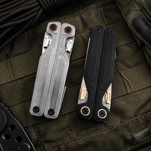 Op de afbeelding: Twee multitools, één zilverkleurig en één zwart, worden getoond op een groene tactische achtergrond. De zilveren tool heeft een geborstelde metalen afwerking, terwijl de zwarte tool een getextureerde grip heeft.