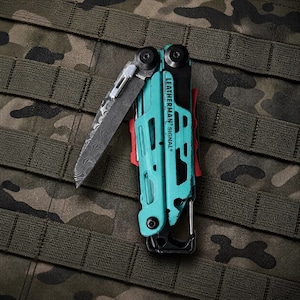 May include: A Leatherman Signal multi-tool featuring a turquoise handle with black accents and a partially deployed Damascus steel blade. The tool includes a carabiner clip at the base and red internal components visible within the frame.