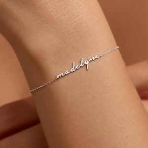 Puede incluir: Una delicada pulsera de plata con el nombre "madelyn" en escritura cursiva. La pulsera es fina y elegante, diseñada para llevar en la muñeca. El nombre es el punto focal, mostrando una joya personalizada.
