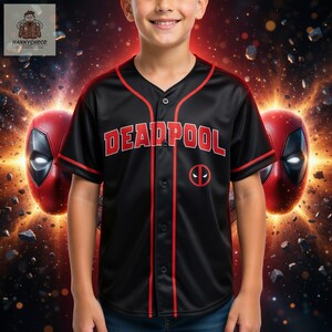 A young boy wearing a black baseball jersey with the word "DEADPOOL" printed on it, standing in front of a background with two red masks and a fiery explosion.