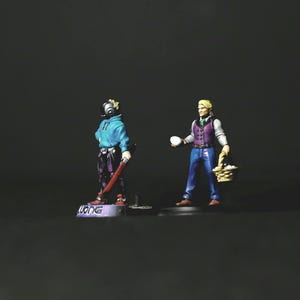 May include: Miniature figurines of two figures. One wears a gas mask and blue hoodie, holding a red object. The other figure holds an egg and a basket of eggs. The base of the first figure has the word "WONG".