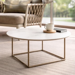 Pode incluir: Uma mesa de centro redonda com um tampo branco com efeito mármore e uma estrutura de metal dourado. Uma caneca branca sobre um descanso de madeira e um livro pequeno estão sobre a mesa. Numa sala de estar.