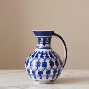 Op de afbeelding: Een wit en blauw keramieken kan met een afgeronde body en een gebogen handvat. De kan is versierd met een herhalend blauw bloemmotief. De rand en het handvat zijn effen blauw.