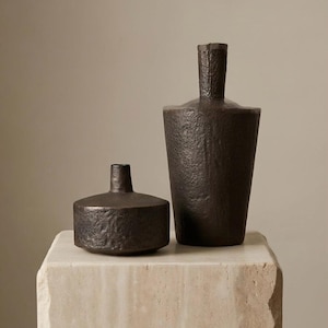 May include: Two dark brown, textured vases of different sizes are displayed on a light-coloured stone block. The taller vase has a cylindrical body and a narrow neck. The smaller vase is round with a short neck. The background is a neutral colour.
