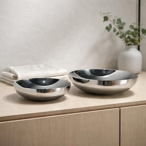 Könnte beinhalten: Zwei Edelstahlschalen in verschiedenen Größen stehen auf einer hellen Holzoberfläche. Die Schalen haben eine glänzende, reflektierende Oberfläche. Ein Stapel weißer gefalteter Handtücher und eine weiße Vase mit Grünzeug befinden sich im Hintergrund.