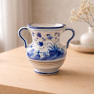 May include: A white ceramic vase with two handles, adorned with blue floral patterns and brown accents. The vase has a wide opening and a rounded base, suitable for decorative purposes.