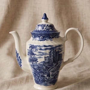 Puede incluir: Una tetera de cerámica color crema con un patrón azul de transferencia que muestra un paisaje de castillo, un asa curva, un pico con detalles florales y una tapa a juego con un remate puntiagudo, decorada con volutas azules.
