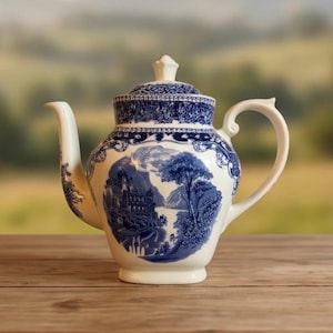 May include: A white ceramic teapot featuring a blue transferware pattern. The design includes a detailed landscape scene with a castle and trees, framed by ornate floral and scrollwork borders on the body, lid, and handle.