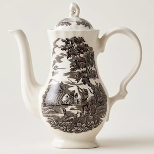 May include: A cream-colored ceramic coffee pot featuring a dark brown transferware print of a rural landscape. The scene depicts a person with a rifle, a dog, a water mill, and trees. The pot includes a matching lid with a finial handle.
