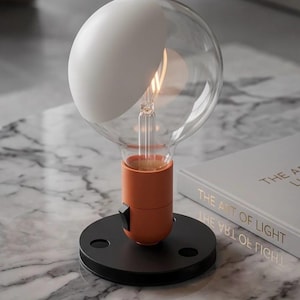 Könnte beinhalten: Eine moderne Tischlampe mit einer großen, klaren Glühbirne und einem weißen, gebogenen Schirm. Die Glühbirne sitzt in einem orangefarbenen Sockel auf einem schwarzen, kreisförmigen Ständer. Ein Buch mit goldener Schrift, "THE ART OF LIGHT", ist sichtbar.