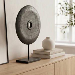 Könnte beinhalten: Ein graues, kreisförmiges Dekorationsobjekt mit einem zentralen Loch, montiert auf einem schwarzen Ständer. Die Skulptur steht auf einem Holzschrank, neben einer kleinen weißen Vase und zwei Büchern. Das Gesamtthema ist Wohnkultur.