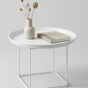 May include: A white side table with a circular tray top and a geometric frame. A small beige vase with dried flowers and a book are on the table. The table is a modern design, suitable for a living room or bedroom.