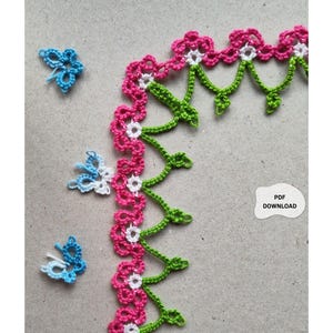 Può includere: Bordura floreale all'uncinetto in rosa, bianco e verde, con tre piccole farfalle all'uncinetto in blu e bianco. La bordura presenta un motivo ripetuto di fiori rosa con centri bianchi e steli verdi. Un piccolo adesivo bianco con la scritta "PDF DOWNLOAD" è nell'immagine.