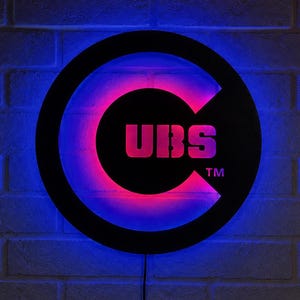 Puede incluir: Un letrero de pared circular que presenta una letra C negra grande con las letras UBS en el centro. El letrero está iluminado con un vibrante brillo LED azul y rosa que resalta el marco circular y el texto central, incluyendo un símbolo TM.