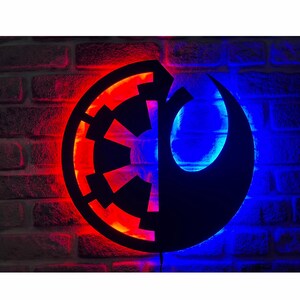 May include: A circular wall sign featuring a split design with the Galactic Empire emblem on the left illuminated in red and the Rebel Alliance symbol on the right illuminated in blue, creating a dual-coloured light effect against the wall.
