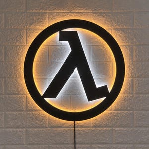 May include: A circular black wall sign featuring the Greek letter lambda in the centre. The sign is illuminated from behind with a bright white light that creates a glowing halo effect against the circular frame and the central symbol.