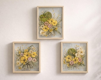 May include: Three framed floral art pieces. Each features a bouquet of yellow daisies, pink flowers, and green foliage. The artwork is set against a light blue background and framed in light wood. The pieces are mounted on a white wall.